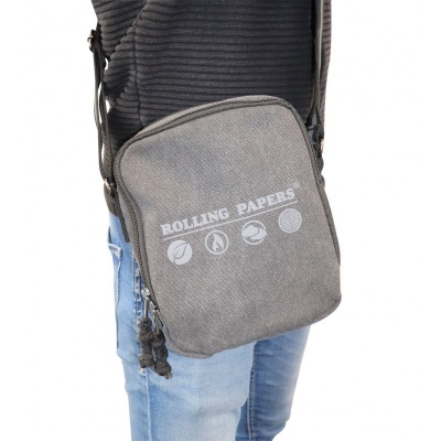 Mochila pequena cinzenta com texto ROLLING PAPERS e ícones em branco, usada por pessoa com jeans e camisola preta.