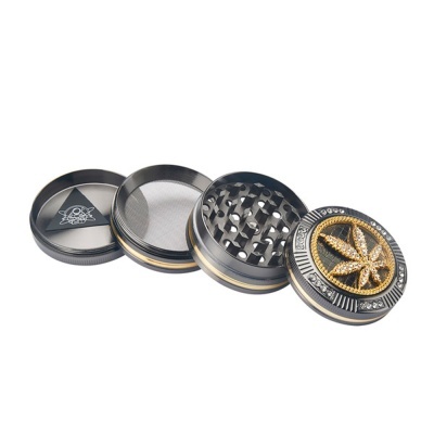 Moedor de metal cinza escuro decorado com folha de cannabis dourada e pedras brilhantes.