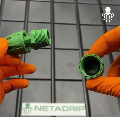 Duas peças de plástico verdes para irrigação, mãos com luvas laranja, fundo de azulejos cinzentos e texto NETADRIP.