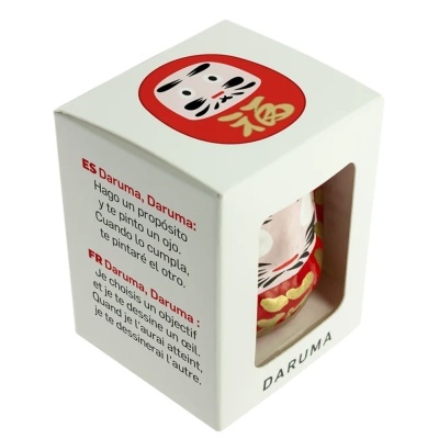 Caixa branca com figura Daruma vermelha dourada e texto explicativo em espanhol e francês
