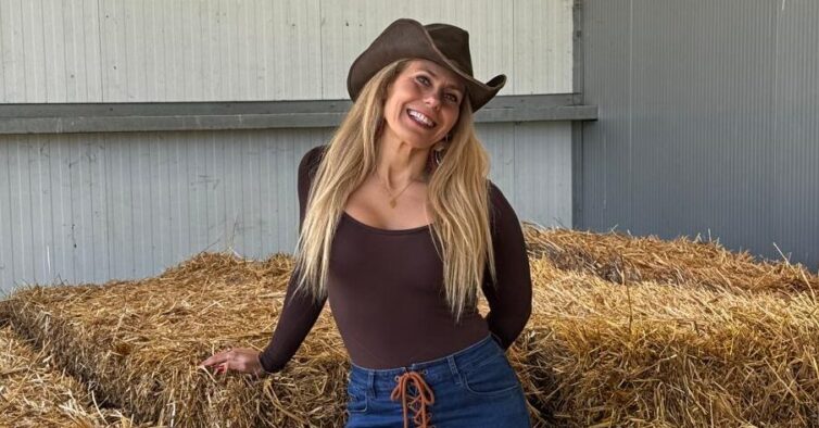 Mulher loira com camisola castanha, calças de ganga e chapéu castanho junto a fardos de palha