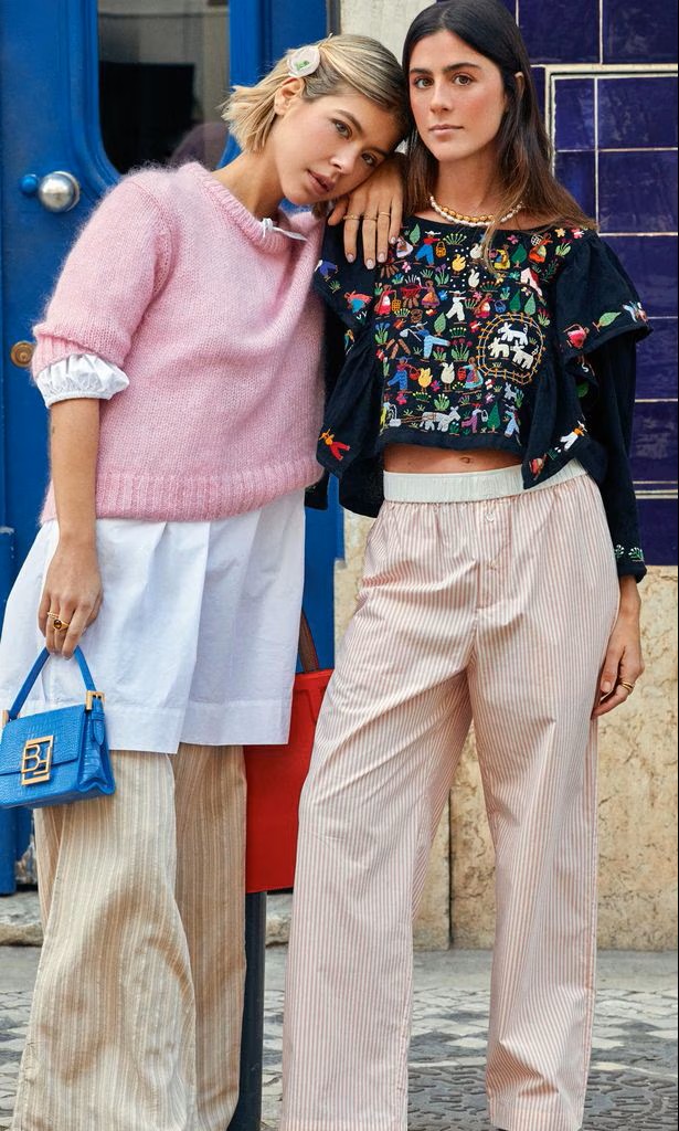 Duas mulheres com roupas casuais coloridas, calças largas às riscas, top bordado e camisola rosa, em ambiente urbano exterior