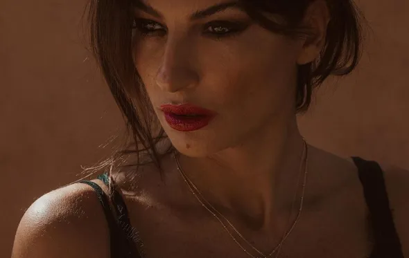 Mulher com maquilhagem escura, batom vermelho, alça preta e colares dourados com fundo terracota