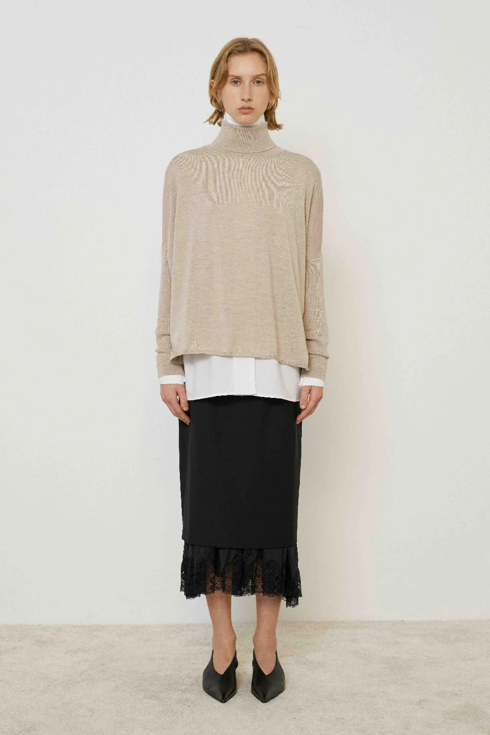 Midi Wool Skirt with Lace Hem Mulher com camisola bege de gola alta, camisa branca, saia preta com renda e sapatos pretos.