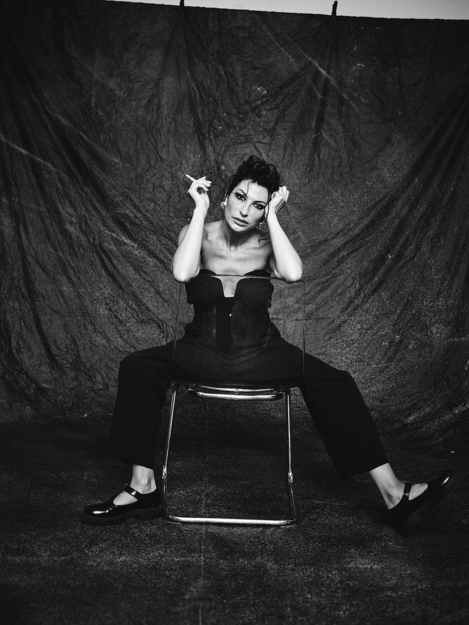 Mulher em roupa preta sentada em cadeira com fundo escuro.