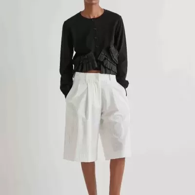 Modelo com casaco preto, bermuda branca de riscas e sapatos pretos em fundo branco