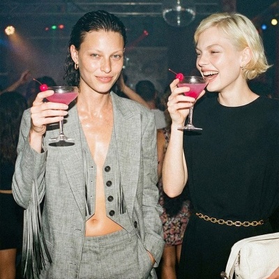 Duas mulheres em discoteca seguram copos de cocktail rosa, uma com fato cinza com franjas e outra com vestido preto e mala branca.