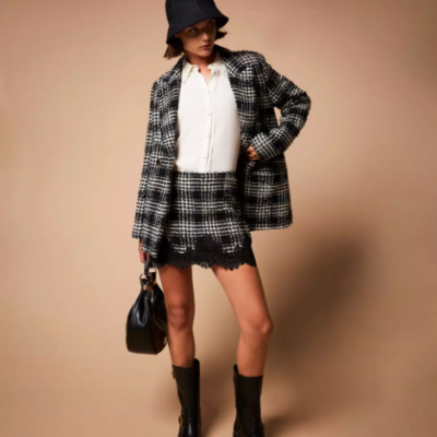 Mulher com roupa xadrez preta e branca, camisa branca, botas pretas e chapéu preto segurando mala preta