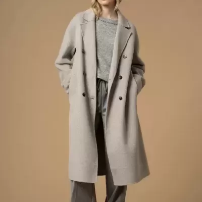Mulher com casaco comprido bege, calças largas cinzentas, camisola cinzenta e sapatos pretos