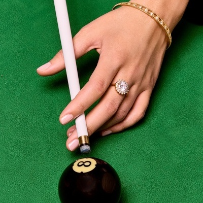 Mão feminina com pulseira e anel segurando taco de bilhar ao lado de bola preta número 8 sobre mesa verde