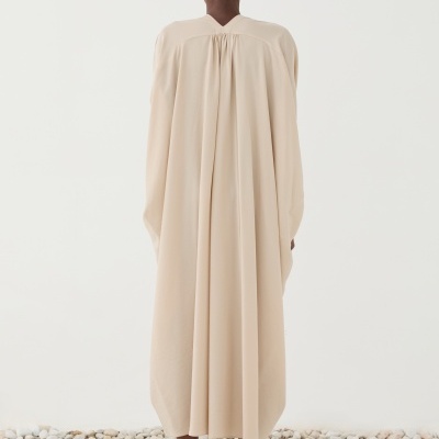 Modelo de costas com túnica bege claro e sandálias castanhas sobre pedras brancas e bege.