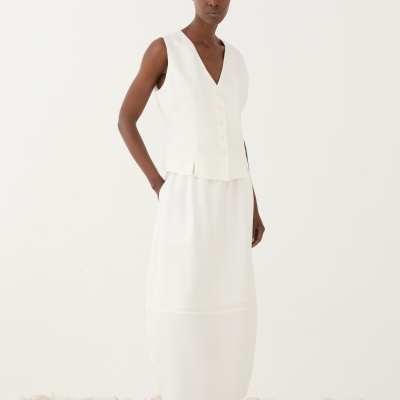 Mulher com conjunto branco de colete e saia, calçada com sapatos brancos, em fundo branco com pedras.