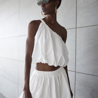 Mulher vestindo conjunto branco com top assimétrico e saia rodada em fundo de parede cinza