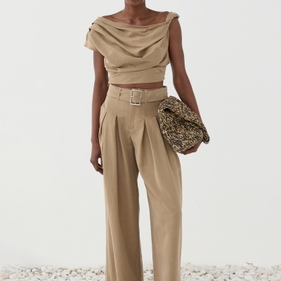 Mulher com roupa bege, bolsa trançada e sandálias castanhas em fundo neutro com pedras no chão