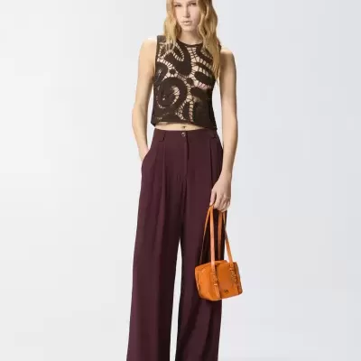 Mulher com roupa de moda casual com top rendado castanho, calças largas castanhas escuras e mala de mão laranja.