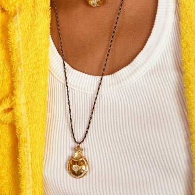 Colar dourado com pendente em forma de frasco e corrente preta com fio dourado sobre roupa branca canelada e casaco amarelo felpudo