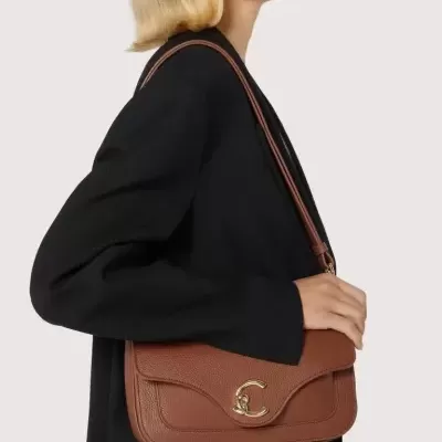 Mala castanha de pele com fecho dourado em forma de C sobre ombro de mulher com casaco preto