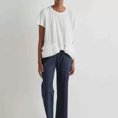 Modelo vestido com t-shirt branca com folhos, calças azul escuro e sapatos pretos em fundo branco.