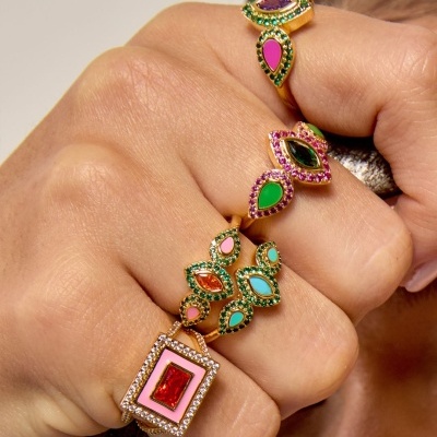Mãos com quatro anéis dourados com pedras coloridas e um anel quadrado vermelho com detalhes