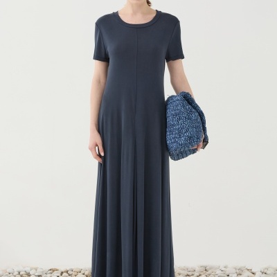 Mulher com vestido comprido azul escuro, bolsa azul trançada e sapatos pretos em chão de pedras brancas