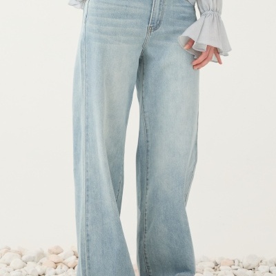 Calças largas de ganga azul clara usadas com blusa branca e sandálias sobre pedras.