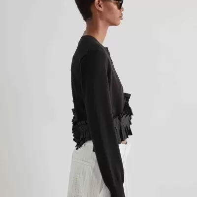 Pessoa de perfil com camisola preta com pregas e calças brancas com riscas verticais