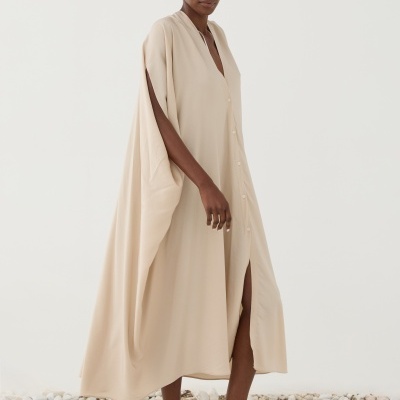 Vestido longo bege claro com botões frontais, tecido liso e leve, usado por modelo de pé sobre pedras brancas.
