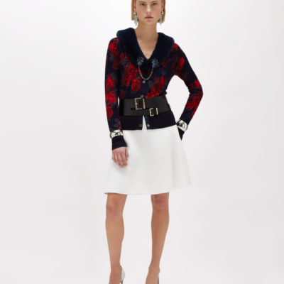 Mulher com casaco azul com flores vermelhas, saia branca e sapatos prateados num fundo branco