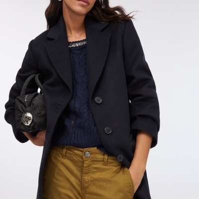 Mulher vestindo casaco negro, camisola de malha azul escura, calças amarelo mostarda e segurando mala preta pequena