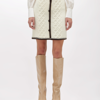 Saia curta de malha creme com padrão trança e bordas pretas, botões pretos, camisa branca e botas bege altas.