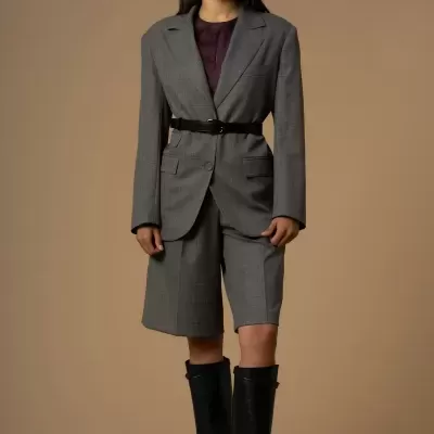 Modelo feminina a usar blazer cinzento xadrez com cinto preto, camisola roxa, bermuda cinzenta e botas pretas altas, em fundo castanho.