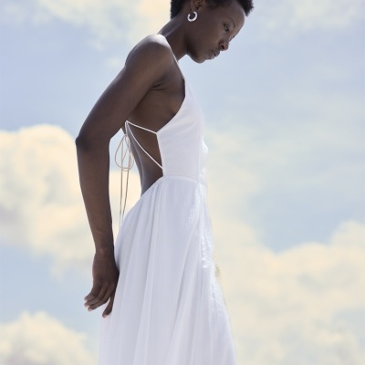 Mulher com vestido branco comprido e costas abertas com tiras finas, fundo de céu azul com nuvens.