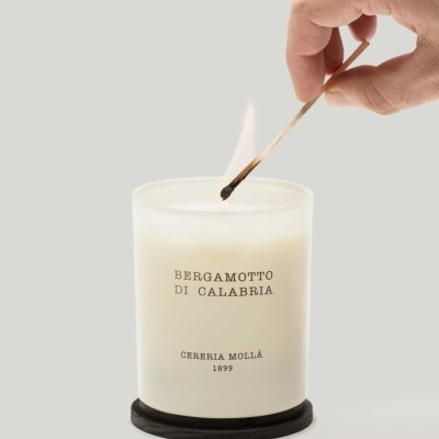 Vela aromática branca em copo de vidro fosco com texto em preto e mão acendendo com fósforo