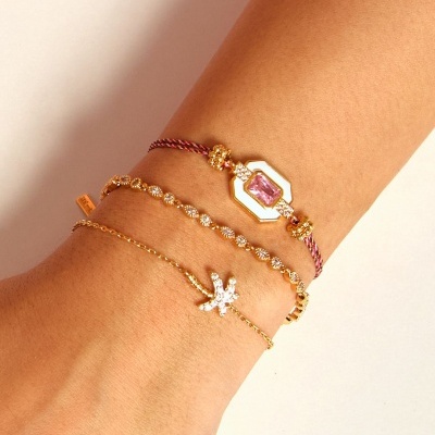 Três pulseiras douradas femininas com pedras e um emblema branco e rosa no pulso.
