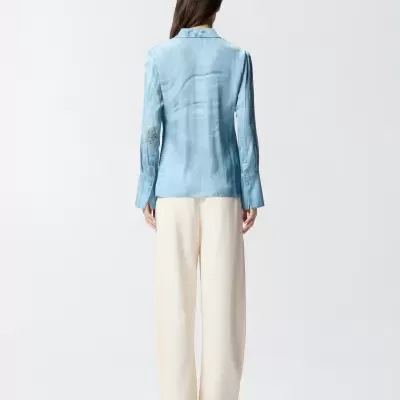 Camisa azul clara feminina com bordado na manga e calças brancas largas vista de costas.