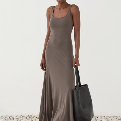 Mulher com vestido longo castanho, sandálias castanhas e bolsa preta em superfície de pedras brancas.