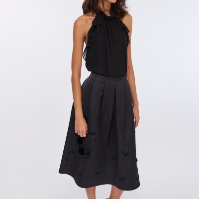 Mulher com blusa preta sem mangas com babados, saia preta midi com aplicações pretas e sapatos de salto alto pretos.