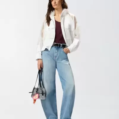 Mulher vestida com camisa branca transparente, camisola vinho, calças de ganga largas, sapatos pretos e mala pequena cinza.