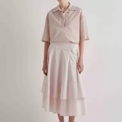 Mulher com camisa rosa clara de manga curta e saia midi rosa clara com folhos e pregas, sapatos cinzentos com laço.