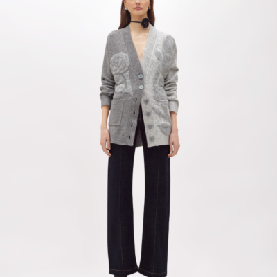 Mulher vestindo casaco cinzento com padrão floral, calças pretas largas e botas pretas em fundo branco.