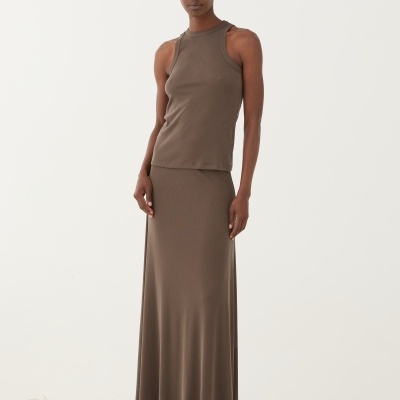 Modelo feminina com conjunto castanho composto por top sem mangas e saia comprida, calça sandálias pretas de plataforma, fundo com pedras.