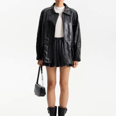 Mulher com casaco preto brilhante, t-shirt branca, saia preta, botas pretas e mala pequena preta com alça de corrente