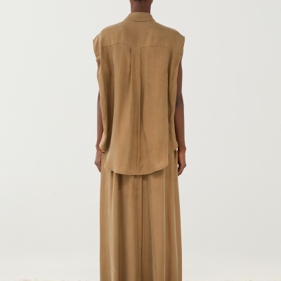 Pessoa de costas com colete e saia castanhos claros em chão de pedras brancas.