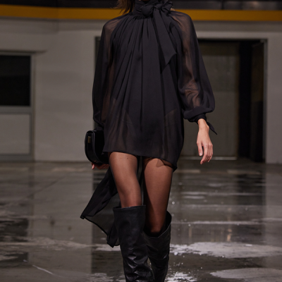 Modelo feminina de vestido preto de gola alta com laço e botas negras altas