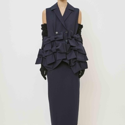 Modelo vestindo vestido longo e colete com laços decorativos em fundo branco.