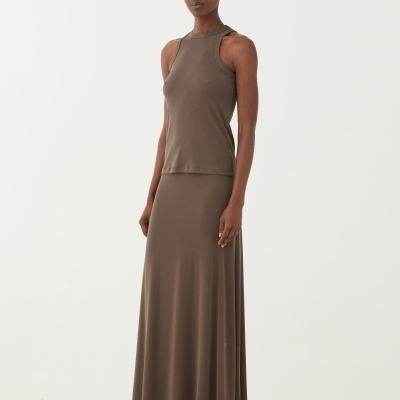 Mulher vestindo conjunto castanho claro com top sem mangas e saia longa, com sandálias pretas, em chão de pedras brancas.