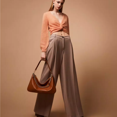 Mulher com cardigan coral de malha e calças largas taupe segurando mala castanha com corrente