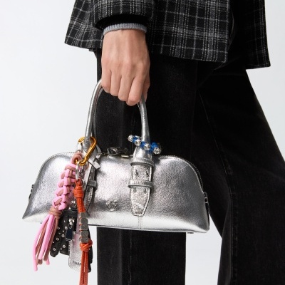 Mala pequena prateada com acessórios coloridos pendurados, segurada por pessoa com casaco xadrez e calças pretas