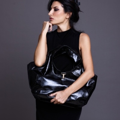 Mulher com vestido preto segurando mala oversized preta de material brilhante com fecho de metal prateado.