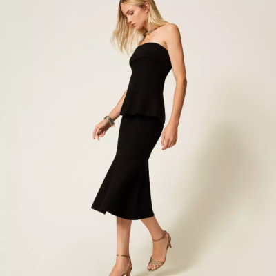 Mulher loira vestindo conjunto preto de top sem alças e saia midi com sandálias douradas.